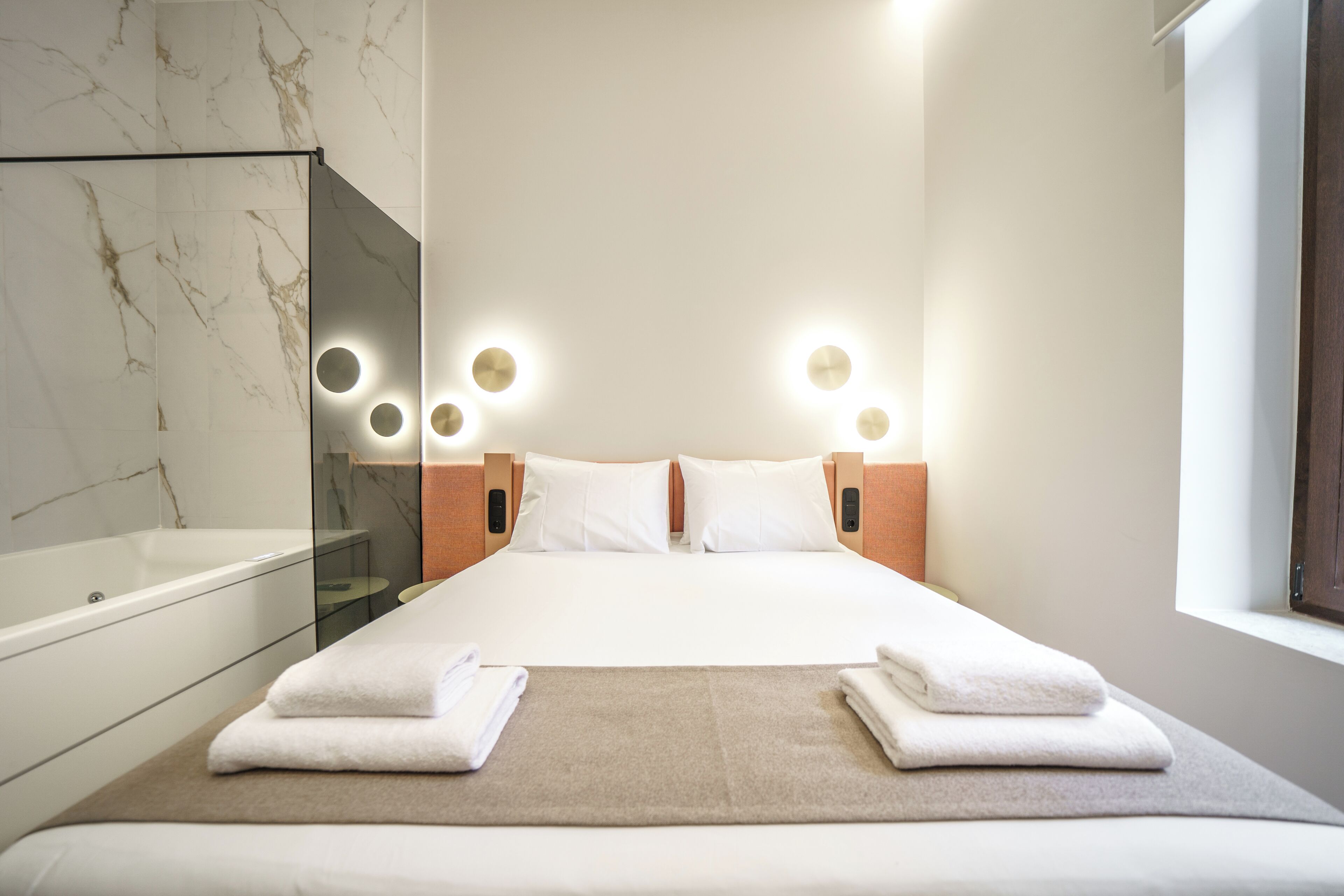 Ett hotellrum med en stor säng, en dusch med glasväggar och utsikt över himlen genom ett fönster.