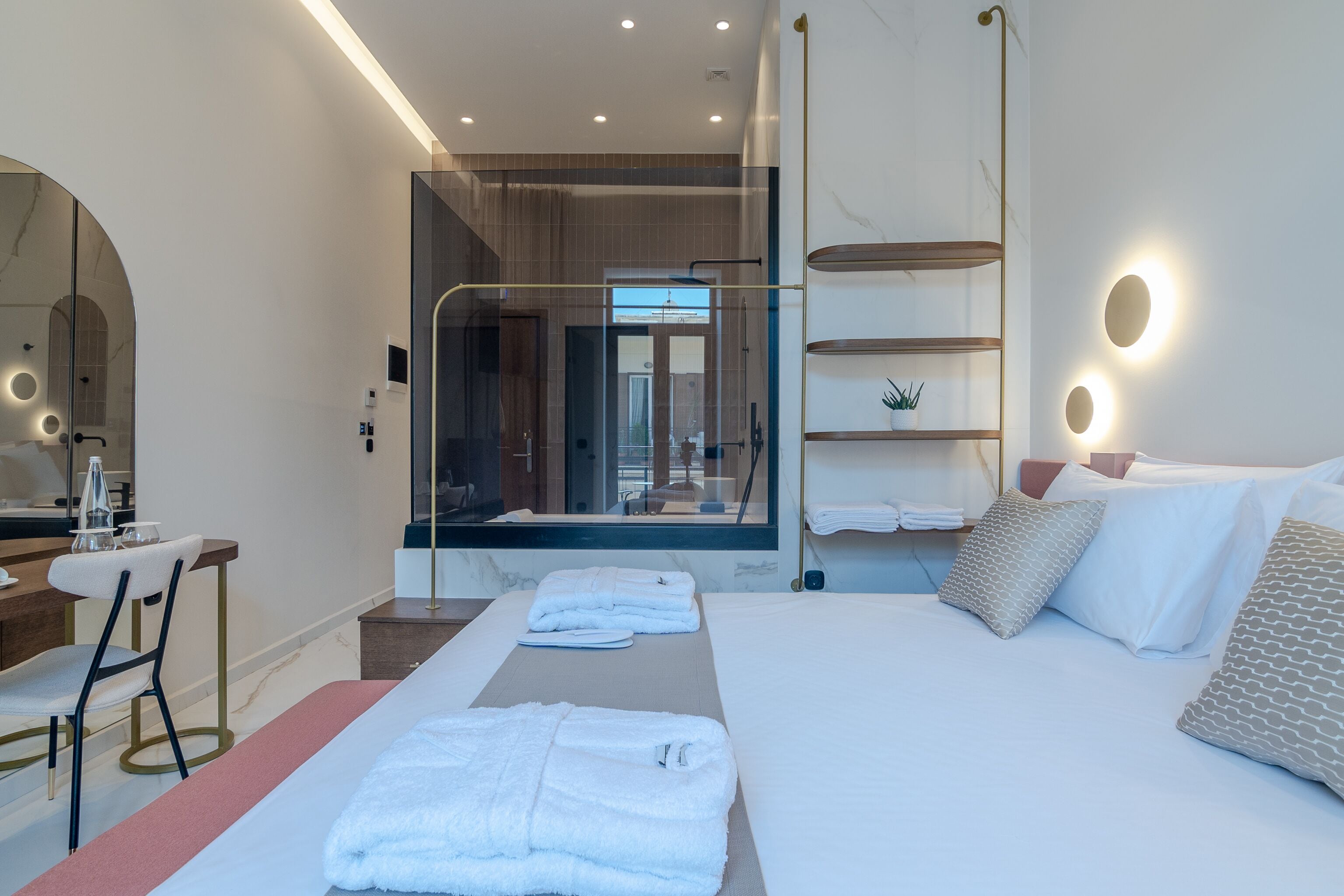 Een moderne hotelkamer met een groot bed, een bureau met stoel, een douchecabine met glazen wanden en een wandplank met handdoeken.