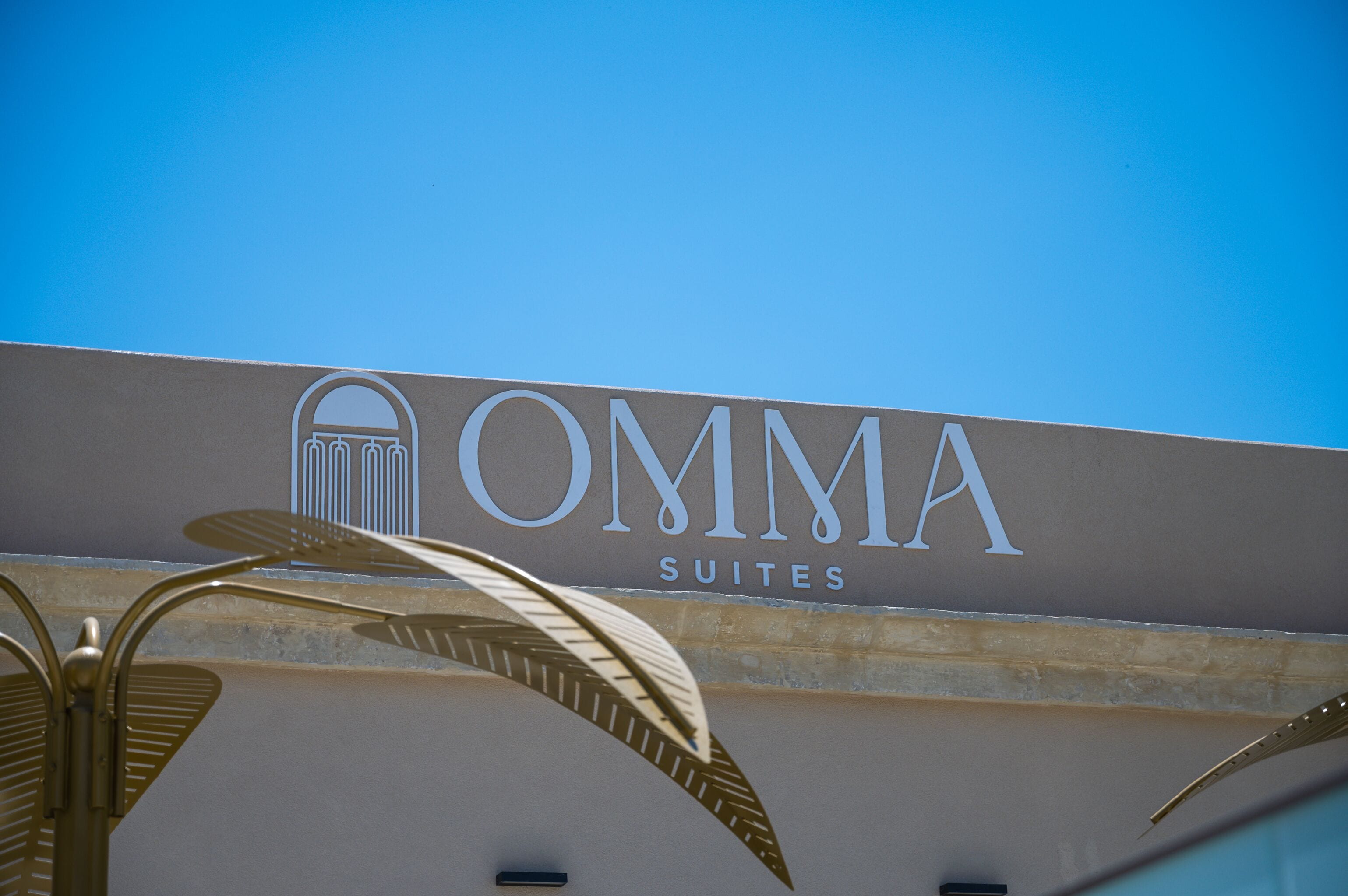 Placa com a inscrição 'OMMA SUITES' e céu azul ao fundo.