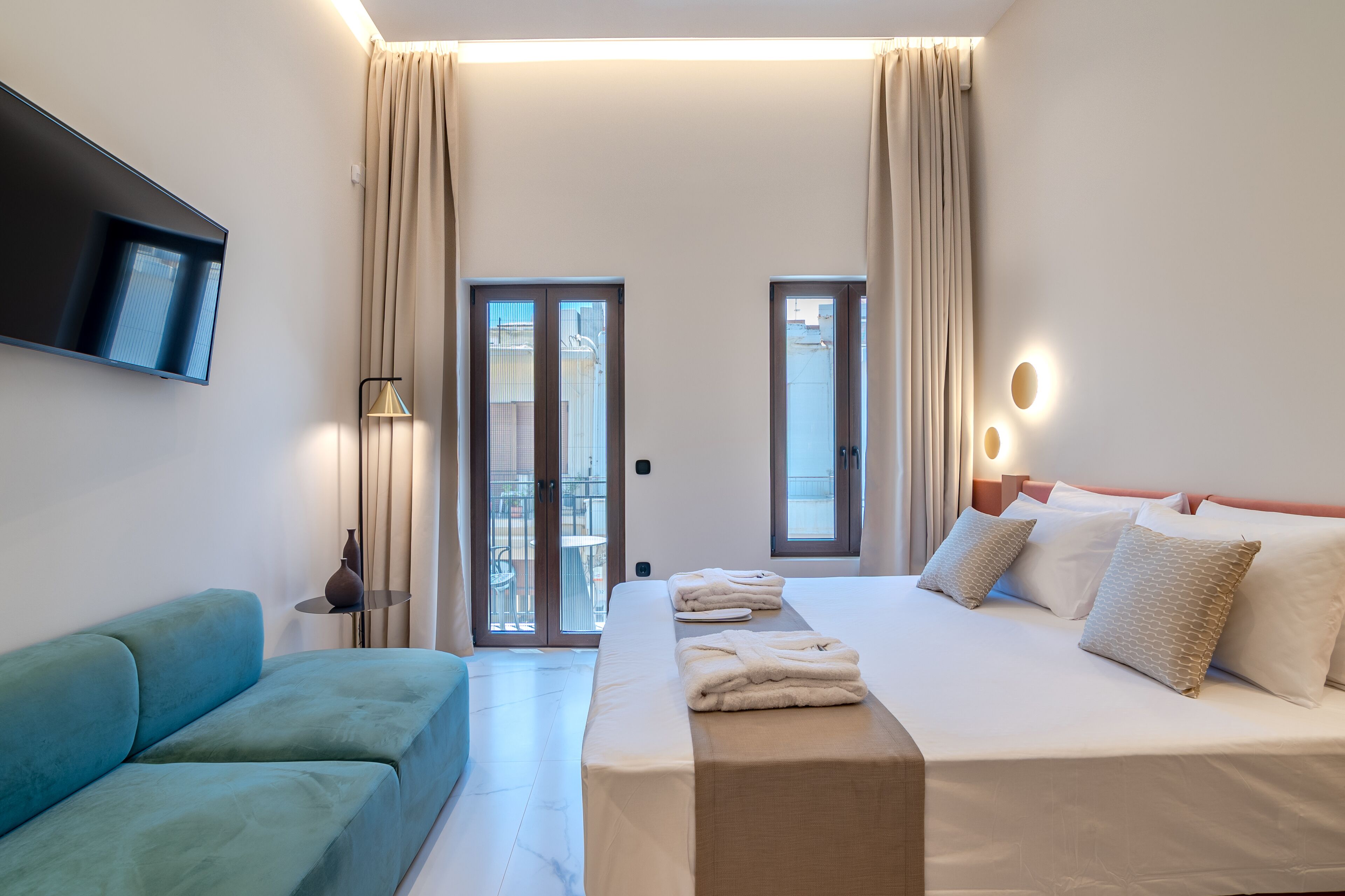 Una habitación de hotel con cama, un sofá azul, un televisor montado en la pared y un balcón con cortinas.
