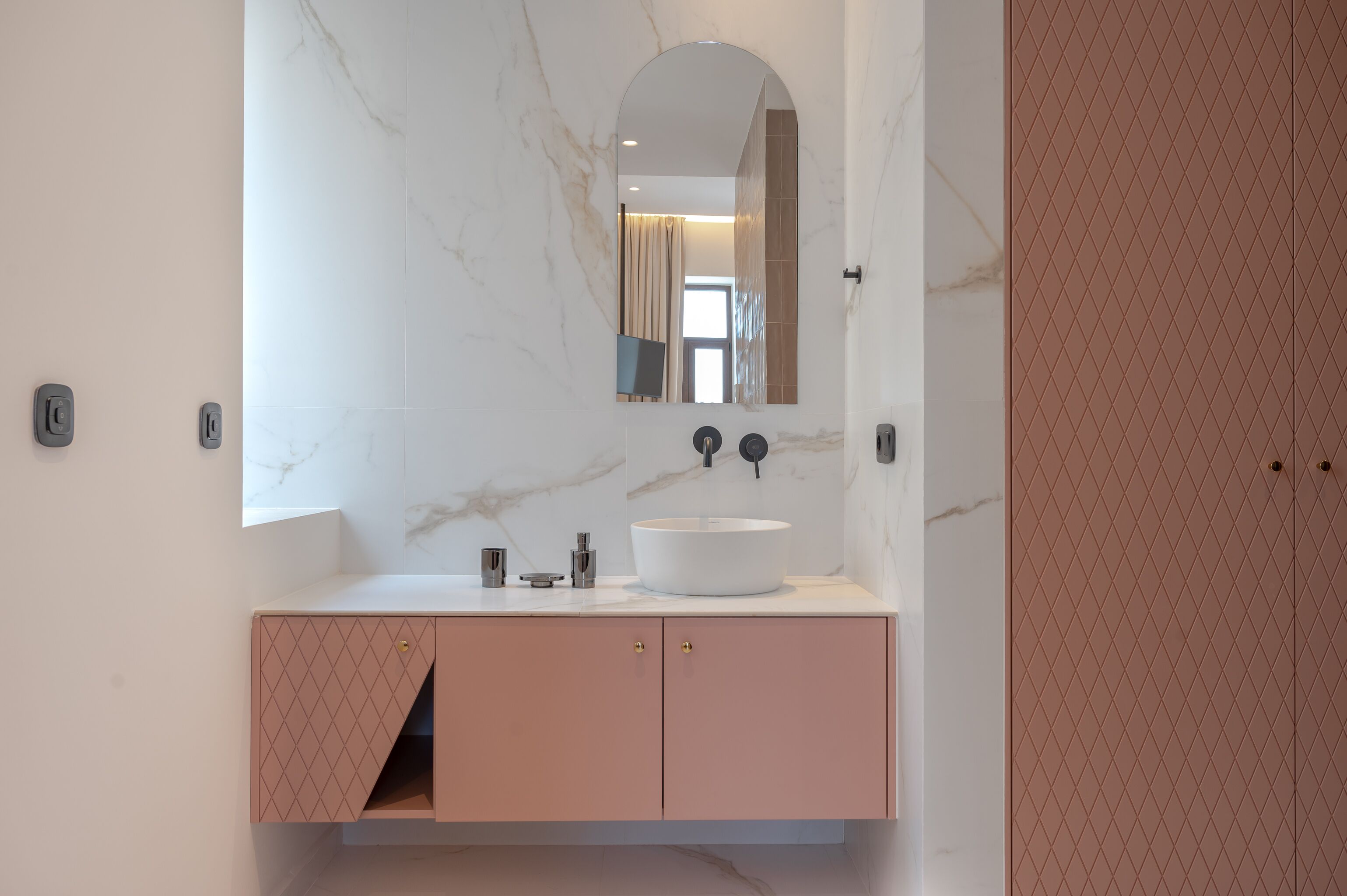 A modern bathroom with a white marble wall, a large mirror, a round sink, and pink cabinets.