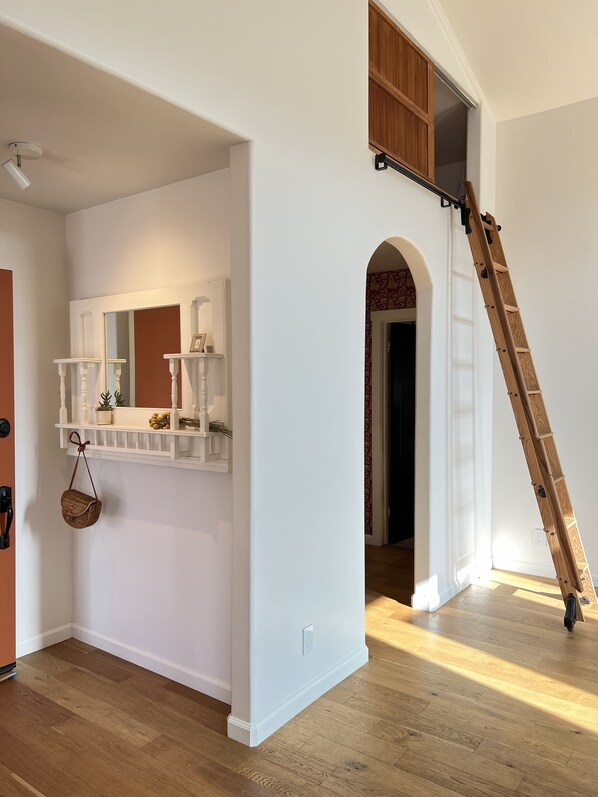 A hallway with a white wall, a wooden floor, a mirror, a small shelf with decorative items, and a wooden ladder leaning against the wall.