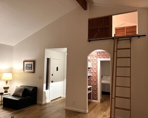 Um quarto com piso de madeira, um sofá de couro preto, uma porta branca e uma escada que leva a um mezanino.