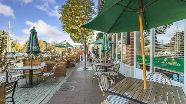 Een terras met houten tafels en stoelen, groene parasols en een bakstenen muur met een glazen etalage waarop de naam van een restaurant staat.