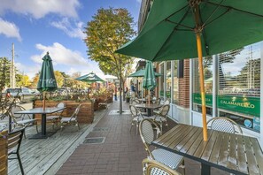 An outdoor cafe with wooden tables and chairs, green umbrellas, and a brick wall with a glass window displaying a restaurant name.