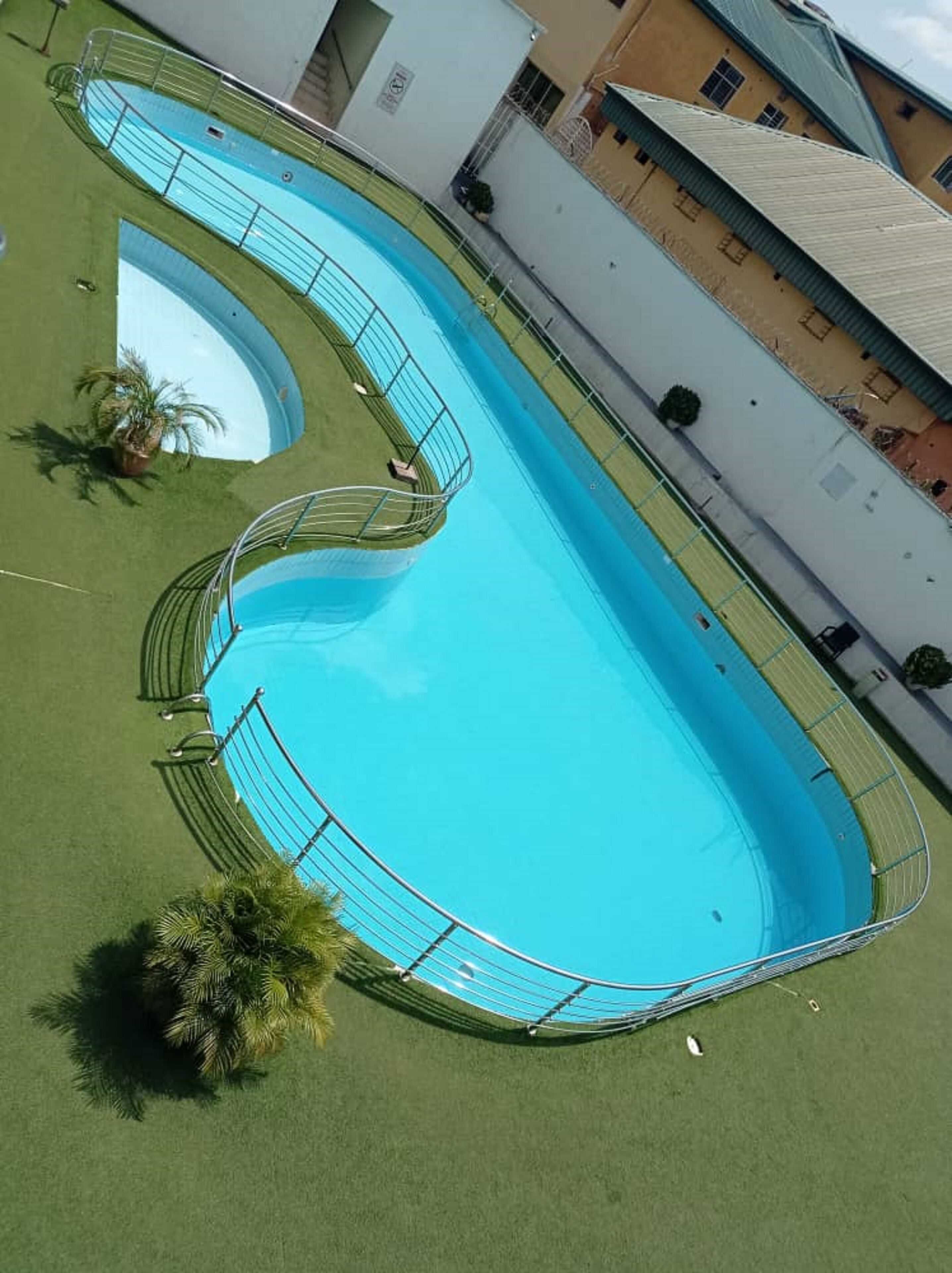 A large, rectangular swimming pool with a curved section, surrounded by a metal fence and a grassy area with palm trees.