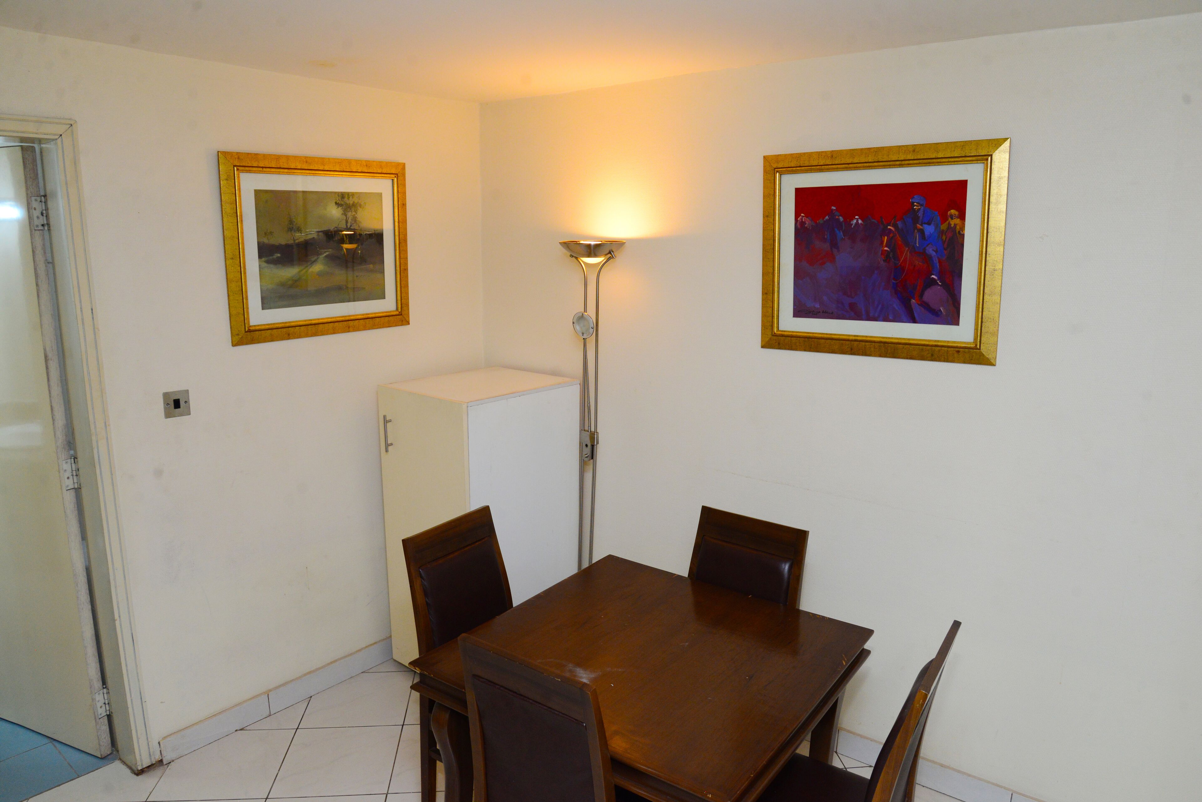 A dining area with a wooden table and four chairs, a floor lamp, and two framed pictures on the walls.
