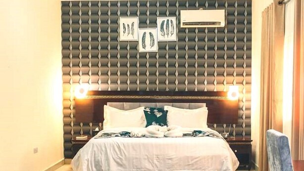 A hotel room with a large bed, a headboard with a textured pattern, two wall-mounted air conditioning units, and framed artwork above the bed. - Golden Royale Hotel (Enugu)