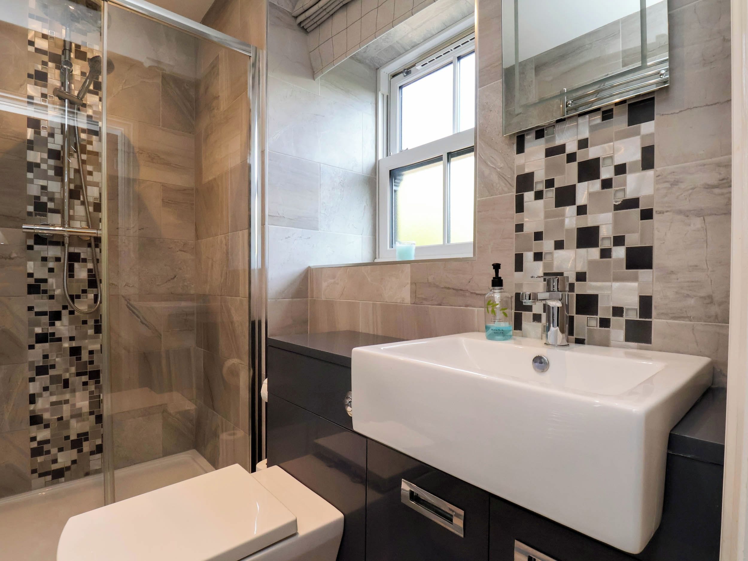 Un bany modern amb dutxa envidriada, un lavabo blanc i un respatller revestit de rajoles.