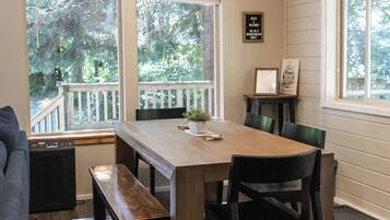A dining area with a wooden table, benches, and chairs. There are windows with blinds, a ceiling light fixture, and a small table with a plant.