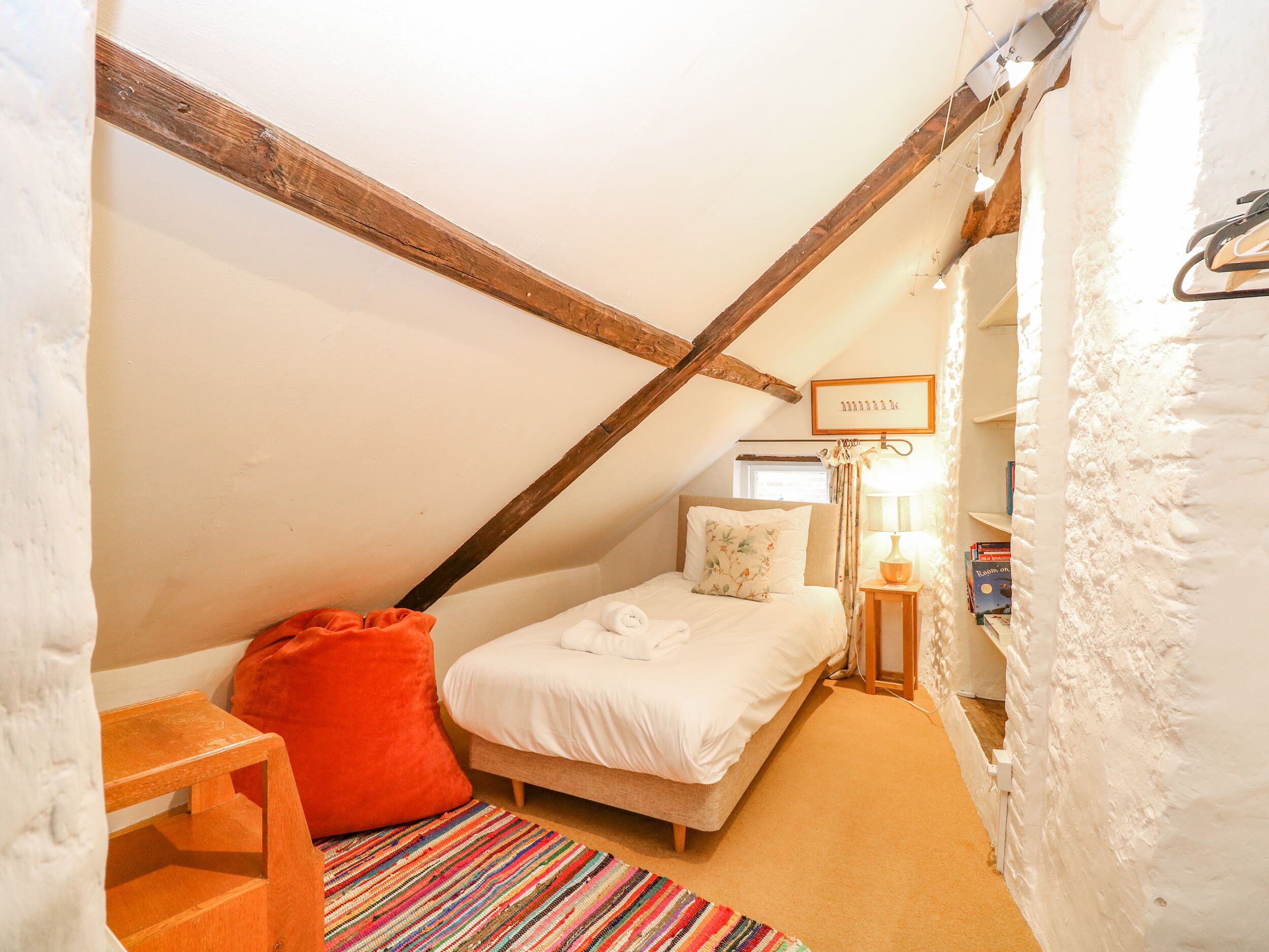 Un dormitorio abrigado en un ático, con techo inclinado, una viga de madera, una cama individual con ropa blanca, un cojín rojo, una mesita, un tapete colorido a rayas, una lámpara de noche y una estantería con libros.