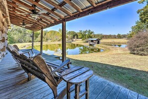 A covered outdoor seating area with wooden chairs and tables, overlooking a pond and a small building in a natural setting. - Unique Loft Cabin: Fire Pit & Fishing Access! (Bells)