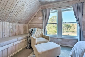 A cozy room with a wooden ceiling, a cushioned bench, a tufted armchair, and a large window with curtains. - Unique Loft Cabin: Fire Pit & Fishing Access! (Bells)