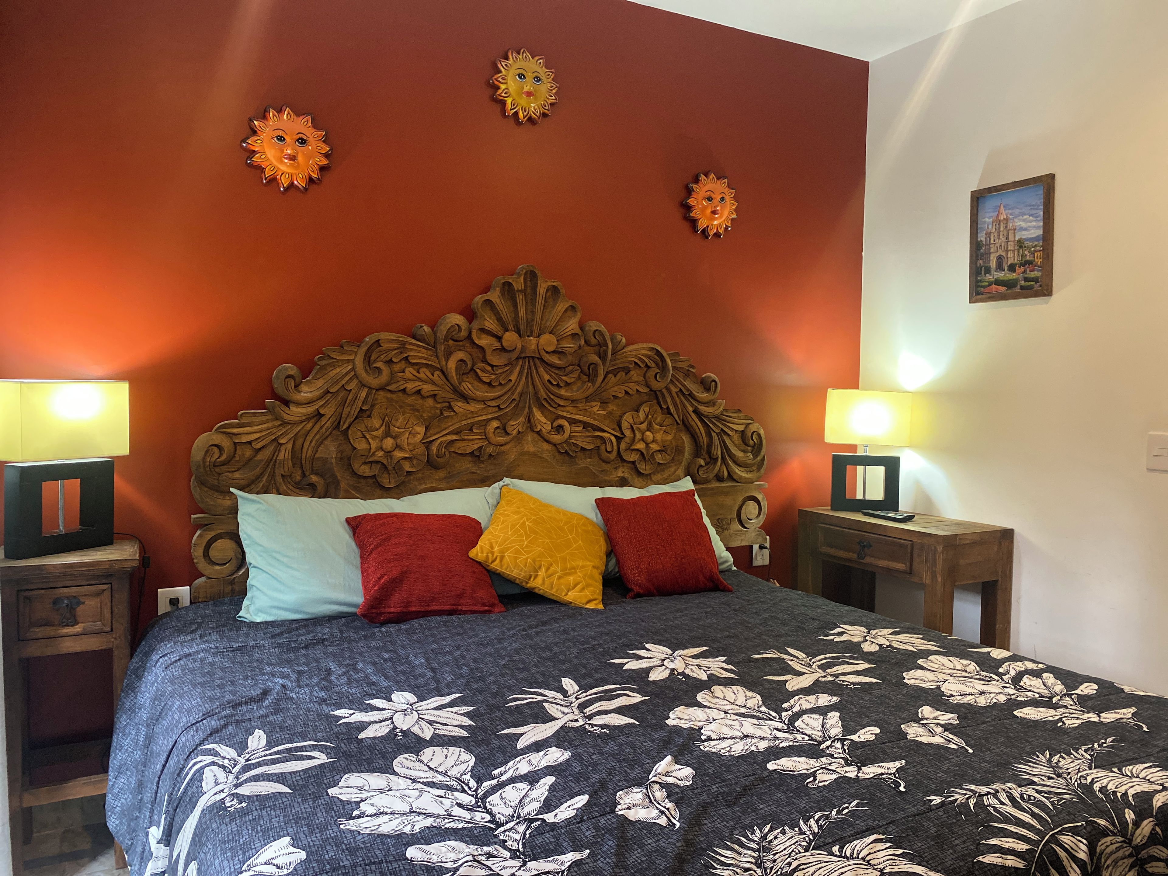 Ein Schlafzimmer mit hohem, hölzernen Kopfteil, vielen Kissen, zwei Nachttischen mit Lampen und einer zudeckenden Bettwäsche mit Blumenmuster.