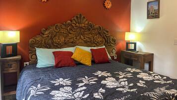 Ein Schlafzimmer mit hohem, hölzernen Kopfteil, vielen Kissen, zwei Nachttischen mit Lampen und einer zudeckenden Bettwäsche mit Blumenmuster.