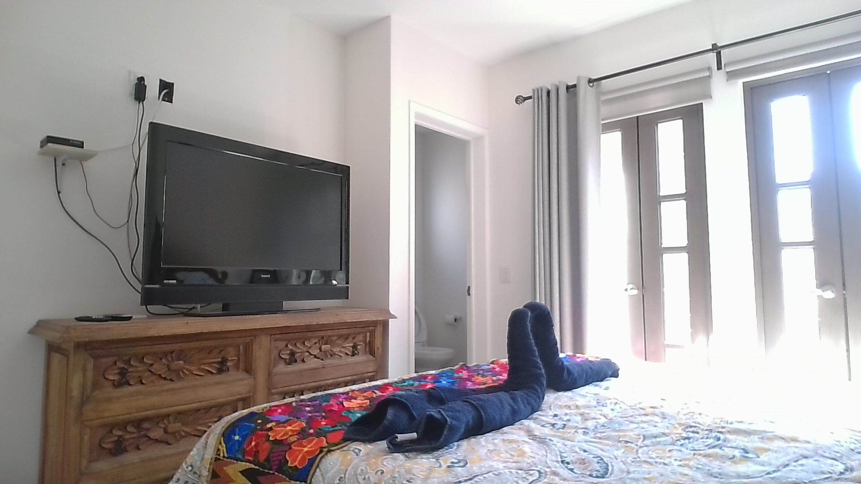 Un dormitorio con una televisión sobre un aparador de madera, una cama con una colcha colorida y una puerta que da a un baño.