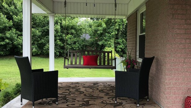 Une véranda couverte avec deux fauteuils en osier, une balançoire et un ventilateur de plafond.