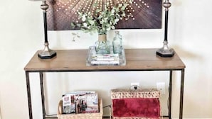 Une console surmontée de deux lampes, un élément décoratif mural, un vase avec des fleurs et des paniers en osier.