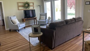 Un salon avec un canapé gris, un fauteuil blanc, un téléviseur posé sur un meuble en bois et une petite table d’appoint avec une lampe.