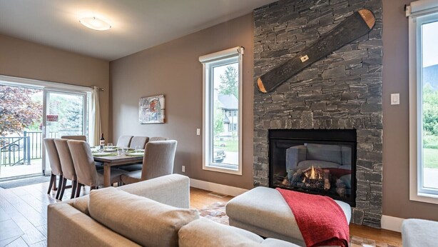 A living room with a stone fireplace, a dining area with a table and chairs, and large windows offering a view of the outdoors. - Aspen Lodge by Revelstoke Vacations (Revelstoke)