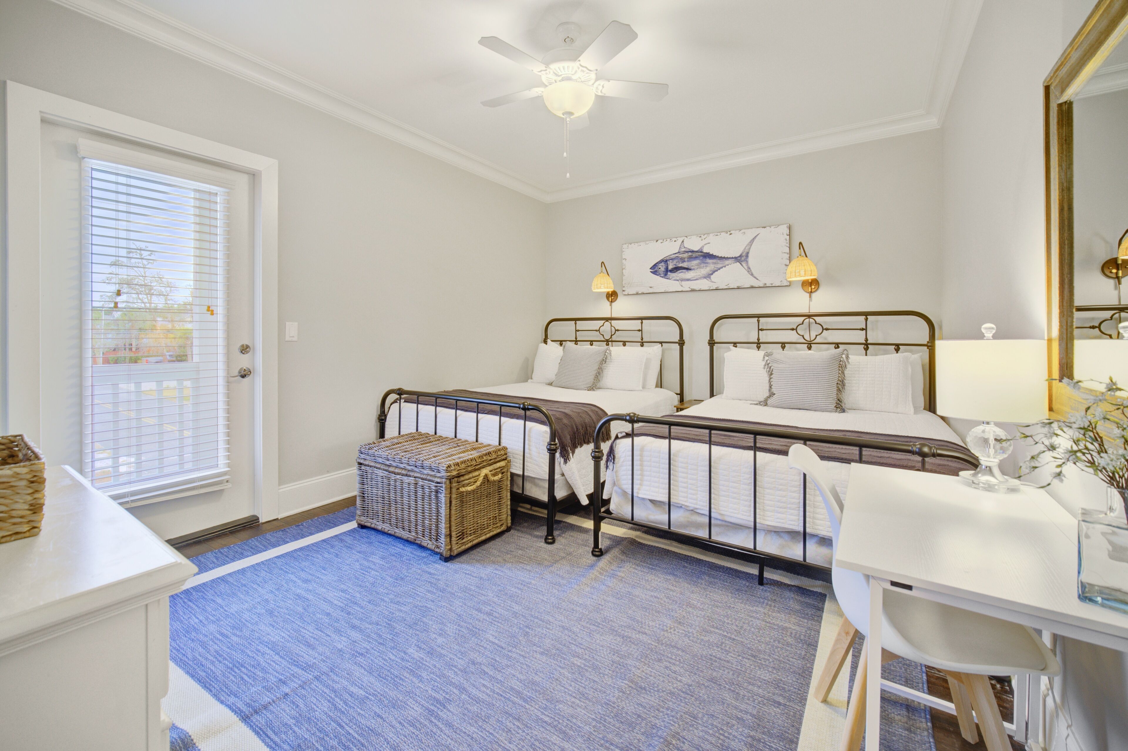 A bedroom with two metal beds, a ceiling fan, a window with blinds, a bedside table, and a wall-mounted lamp.