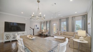 A spacious living room with a wooden dining table, white wicker chairs, a beige sofa, and a TV mounted on a white cabinet. - Steps to 30A - Golf Cart - Kid & Friendly! (Santa Rosa Beach)