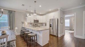 A modern kitchen with white cabinets, stainless steel appliances, and a wooden dining table with bar stools. - Steps to 30A - Golf Cart - Kid & Friendly! (Santa Rosa Beach)