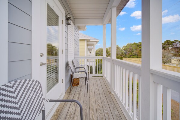 A covered porch with white railings, two chairs, and a view of a yellow house in the background.