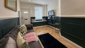 Un salon avec un canapé, un tapis noir, un téléviseur posé sur un meuble et une fenêtre avec des stores.