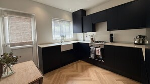 Une cuisine moderne avec des meubles noirs, un plan de travail blanc, une fenêtre avec des stores et un parquet en bois disposé en motif chevron.