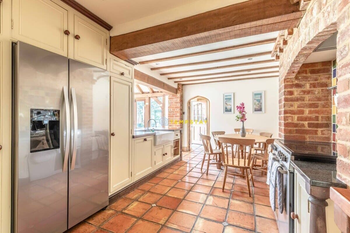 Een keuken met een roestvrijstalen koelkast, houten kastjes, een betegelde vloer, een eethoek met stoelen en een bakstenen muur.