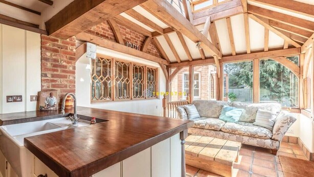 Una cocina con encimera de madera, un lavamanos y una ventana con detalles decorativos. En la zona de estar contigua hay un sofá y una mesa.
