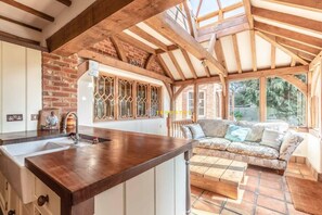 A kitchen with a wooden countertop, a sink, and a window with decorative glass. There is a sofa and a table in the adjacent living area. - 4 Bed Windsor Countryside Escape - By Tempstay (Maidenhead)
