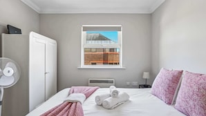 Une chambre d’hôtel avec un lit, une fenêtre donnant sur des immeubles, une armoire, un ventilateur et une table de chevet avec une lampe.