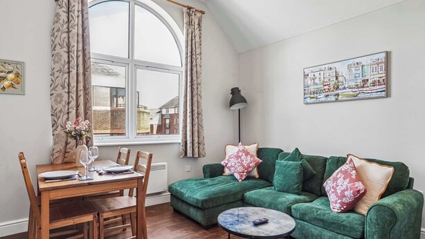 Un salon chaleureux avec un coin repas, une grande fenêtre avec des rideaux, un canapé vert agrémenté de coussins à motifs floraux et un tableau accroché au mur.