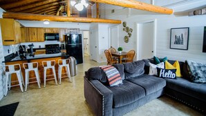 A spacious living area with a dark grey sofa, a wooden dining table with chairs, a kitchen with a black refrigerator, and a high ceiling with exposed beams. - Log Cabin | Guadalupe River | Kayaks | Ping Pong (Seguin)