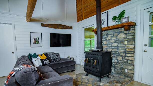 A cozy living room with a stone fireplace, a grey sofa with colorful cushions, and a flat-screen TV on the wall. - Log Cabin | Guadalupe River | Kayaks | Ping Pong (Seguin)