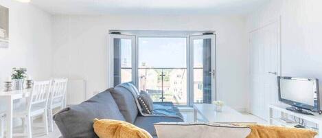 A modern living room with a grey sofa, a TV, and a dining area with white chairs and a table.
