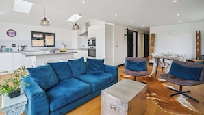 Un soggiorno moderno con un divano blu, un'area pranzo con tavolo e sedie, una cucina con mobili bianchi e un soffitto con faretti.