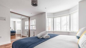 A bedroom with a bed, a window with blinds, a mirror, and a door leading to another room. - 4-Bedroom Retreat in Slough - By Tempstay (Slough)
