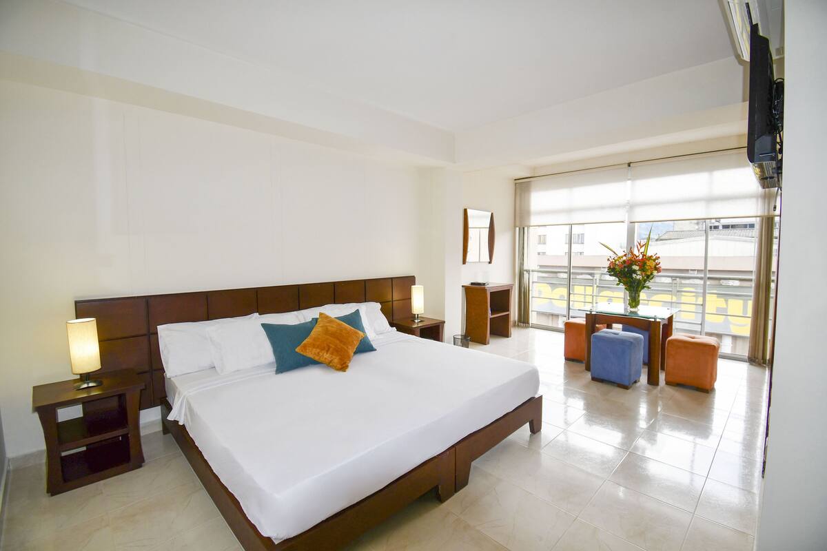 A hotel room with a large bed, a nightstand, a table, and a view of the city through the window.