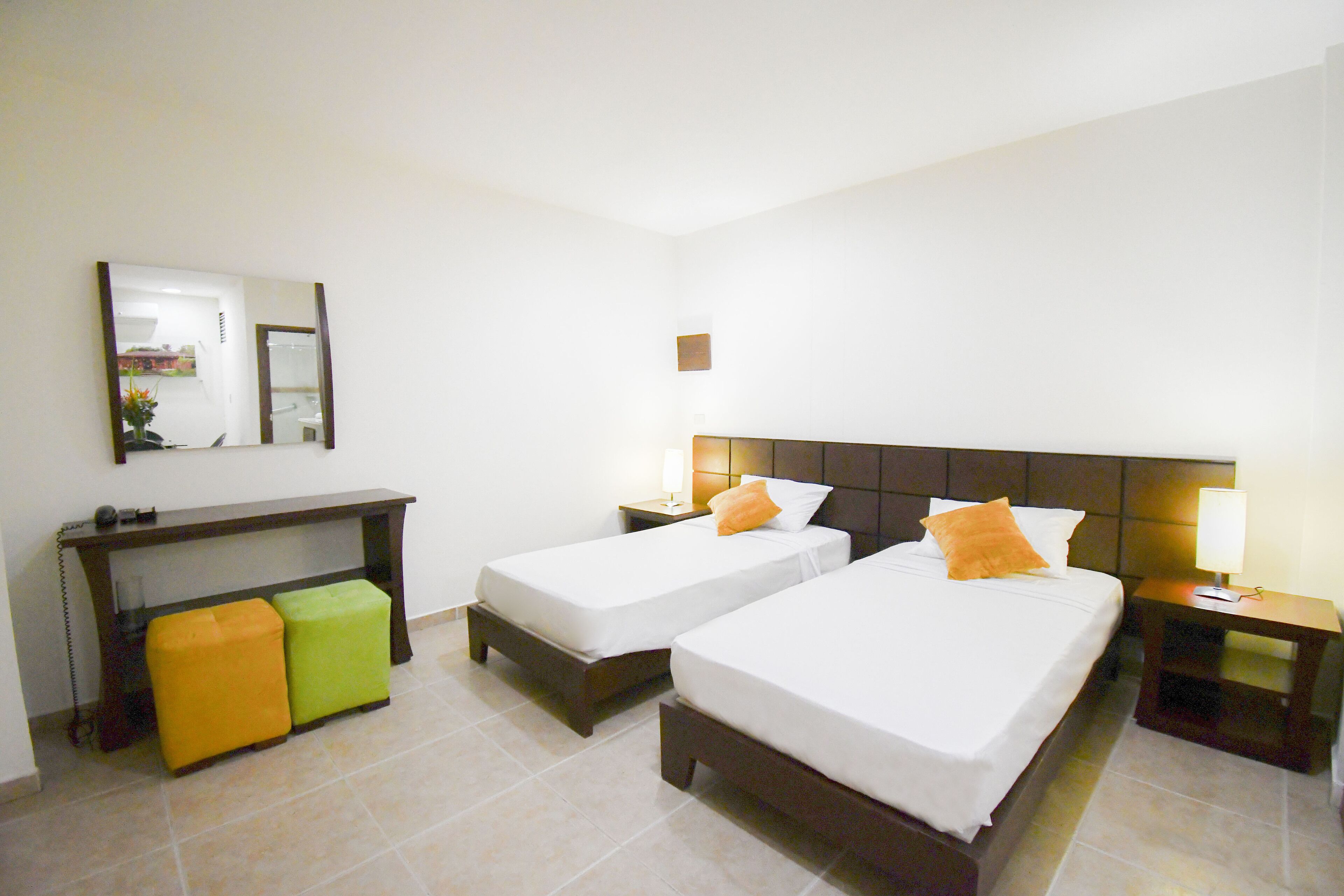 A hotel room with two beds, a nightstand with a lamp, a small table, and a bench.