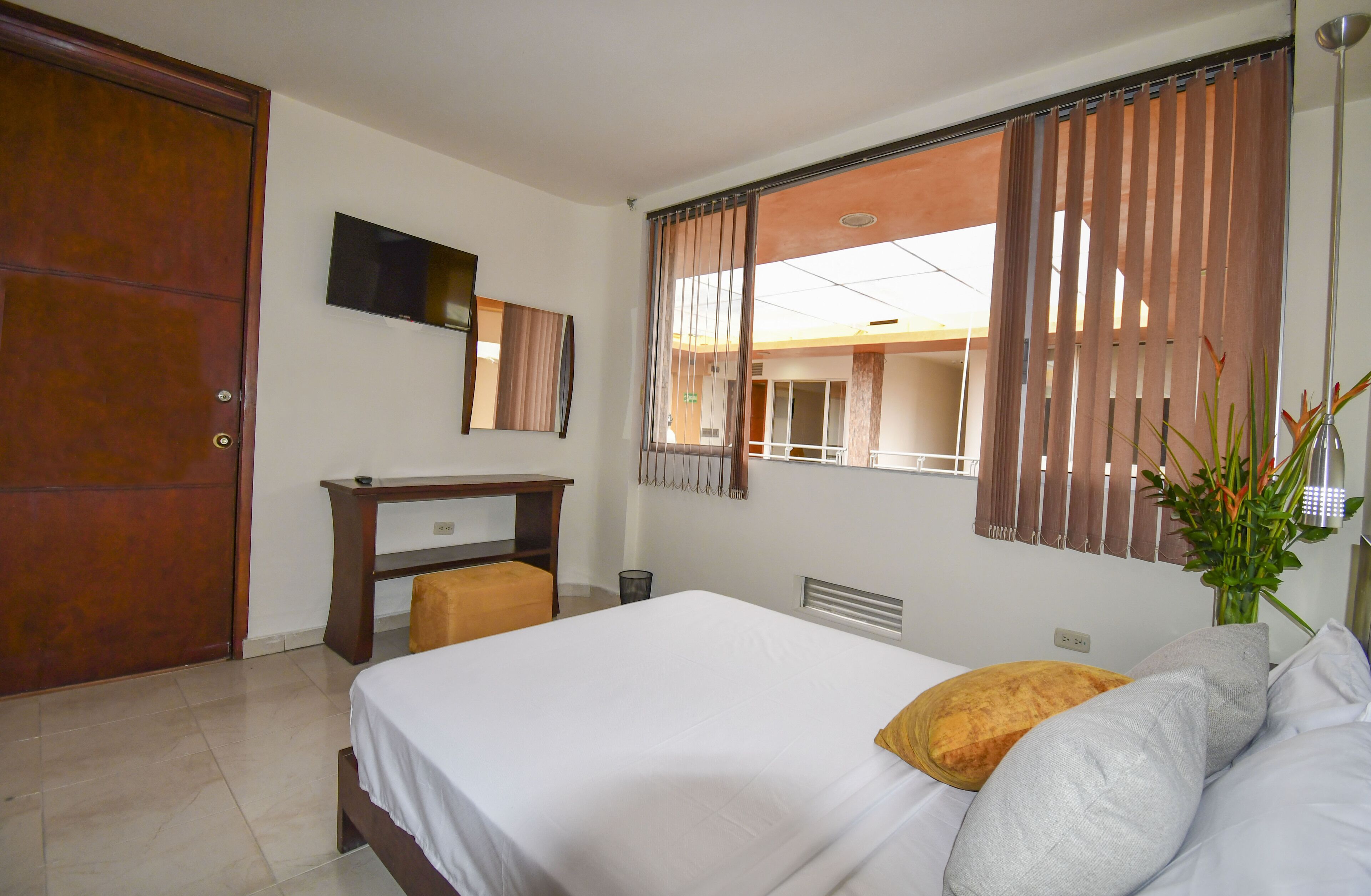 Une chambre d’hôtel avec un lit, un bureau, une chaise, une télévision, une fenêtre avec des rideaux et une plante.