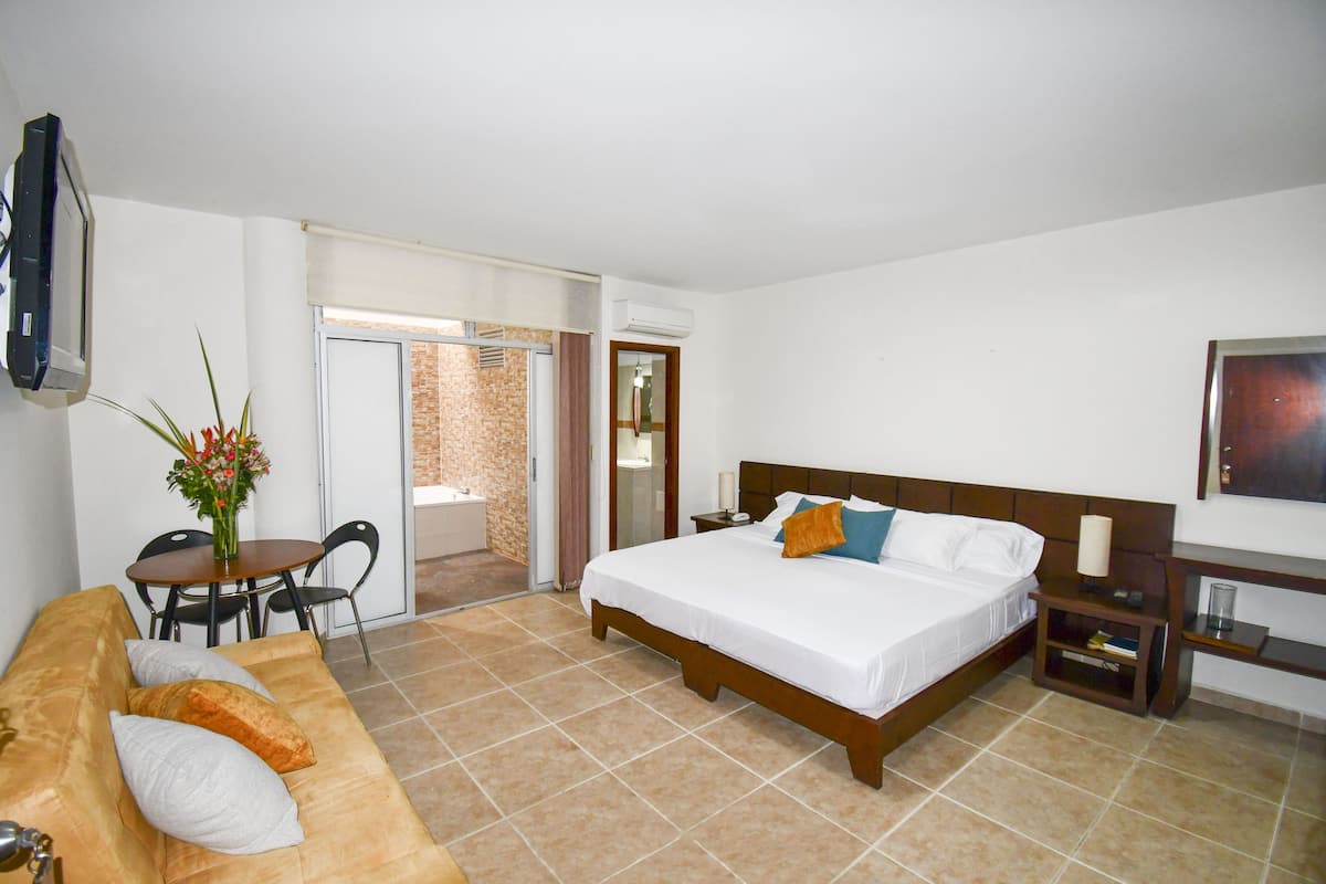 A hotel room with a bed, a sofa, a small table with chairs, a TV, and a door leading to a balcony.