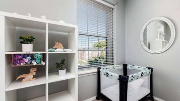 A room with a white crib, a white shelving unit with toys and plants, a window with blinds, and a round mirror on the wall.