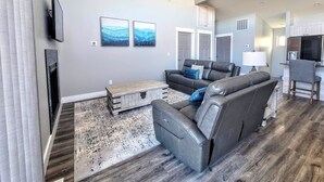 A modern living room with a gray sofa set, a wooden coffee table, and a patterned rug. There are two framed pictures on the wall, a flat-screen TV mounted above the sofa, and a kitchen area visible in the background. - Beautiful Brand New Home with the BEST VIEW OF THE MISSOURI RIVER (Wolf Creek)