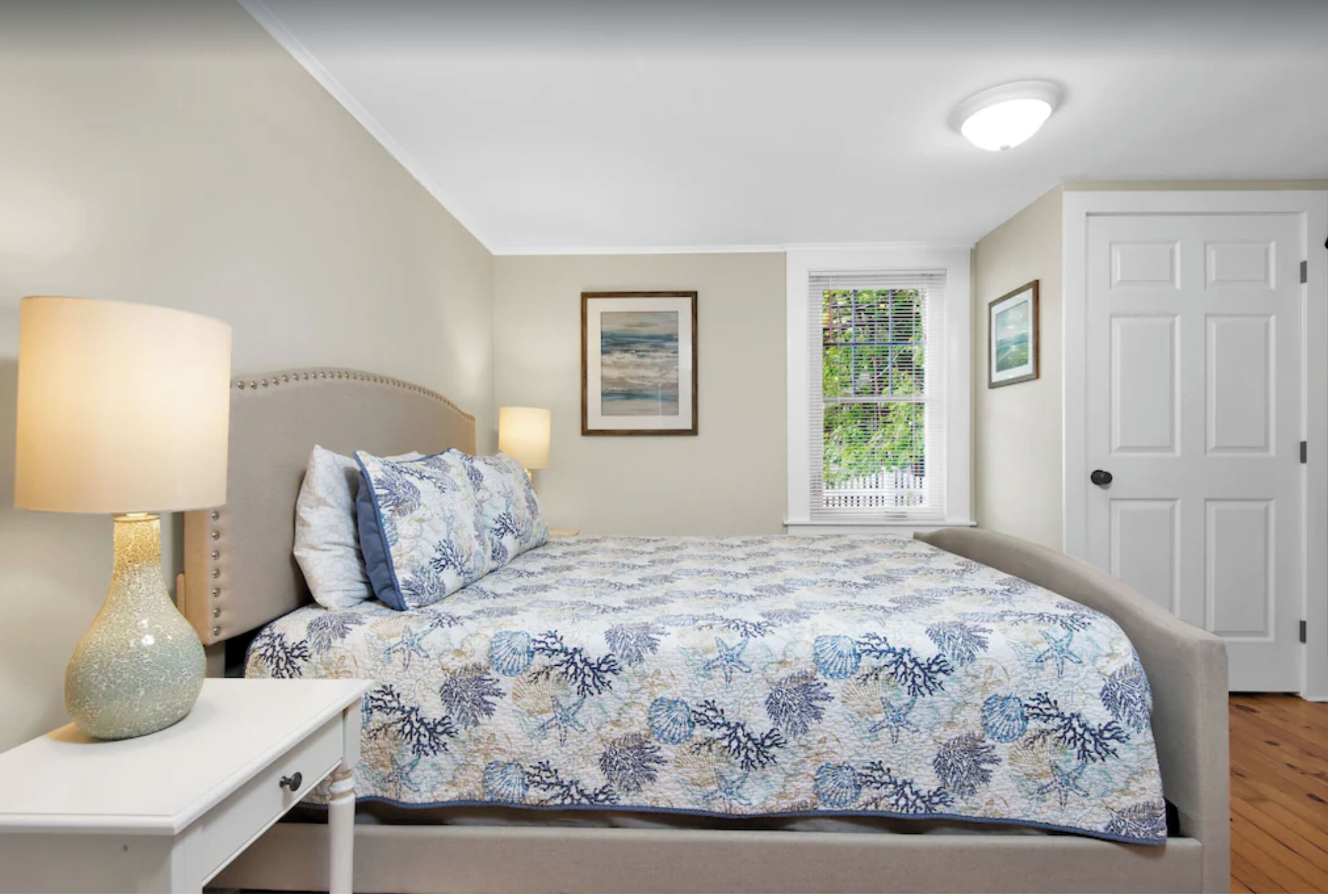Un dormitorio con cama, mesita de noche, lámpara, cuadro enmarcado, ventana con persianas y una puerta.