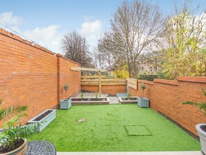 Un giardino a parete di mattoni con erba sintetica, aiuole rialzate e un gazebo in legno.