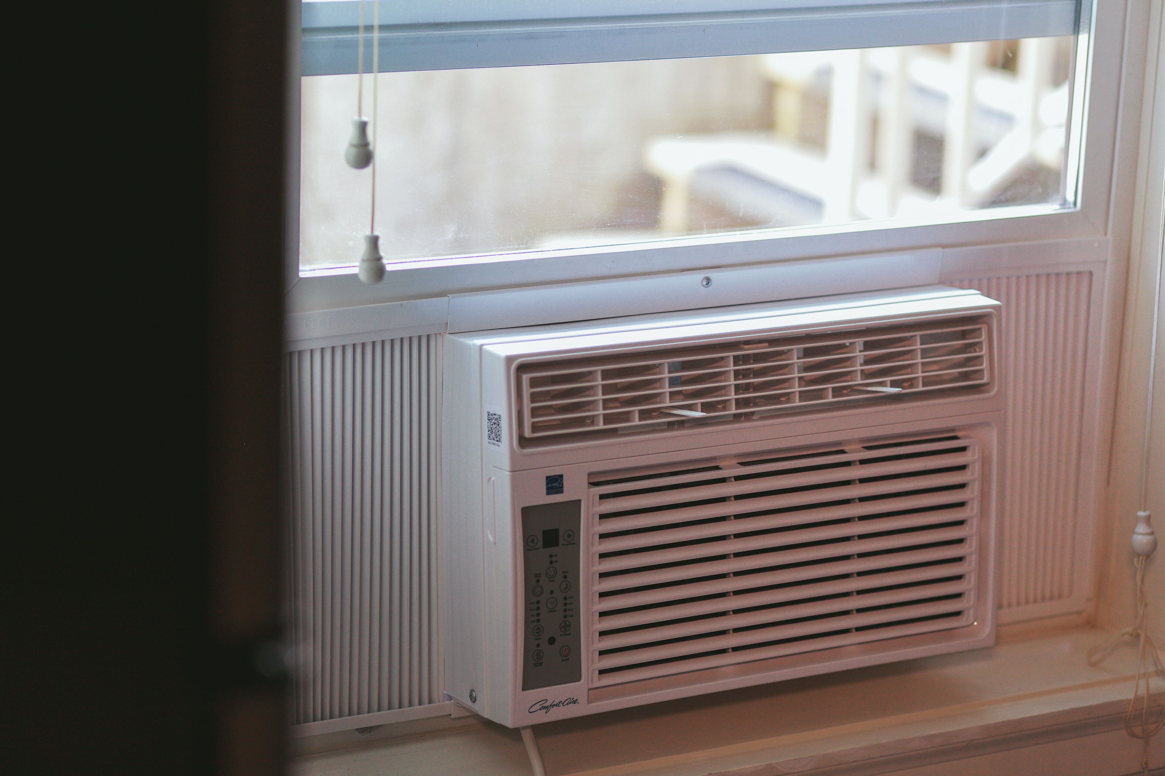 A white wall-mounted air conditioner with a control panel, located near a window with blinds.