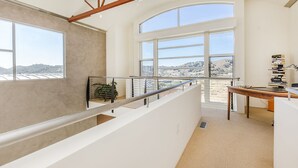 A spacious room with a high ceiling, large windows offering a view of the hills, a staircase with a metal railing, and a desk with books and a lamp. - Ultra modern loft with 3-floor sunlight, 15 min from GGbridge (San Rafael)